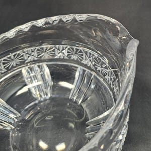 Op de afbeelding: Een creamer van helder glas met een geschulpte rand en een band van ingewikkelde sterpatronen rond de bovenkant. Het onderste gedeelte van het lichaam vertoont verticale, gegroefde panelen die zich uitstrekken naar de basis.