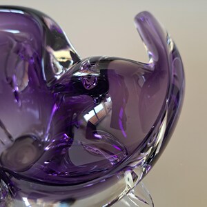 K&ouml;nnte beinhalten: Eine tiefviolette Glasschale mit organischer, asymmetrischer Form. Das durchscheinende Material weist glatte, flie&szlig;ende Kurven und dicke, abgerundete Kanten auf, die das Licht einfangen und so verschiedene Violett- und Dunkellila-T&ouml;ne erzeugen.