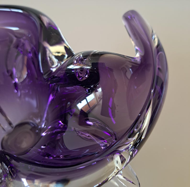 K&ouml;nnte beinhalten: Eine tiefviolette Glasschale mit organischer, asymmetrischer Form. Das durchscheinende Material weist glatte, flie&szlig;ende Kurven und dicke, abgerundete Kanten auf, die das Licht einfangen und so verschiedene Violett- und Dunkellila-T&ouml;ne erzeugen.