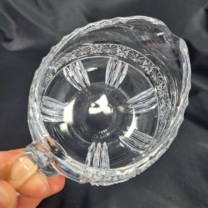Op de afbeelding: Een creamer-schenkkan van helder glas met een decoratief geslepen patroon rond de bovenrand en verticale gegroefde details langs de binnenkant van de bodem. De kan heeft een gebogen handvat en een kleine schenktuit aan de bovenrand.