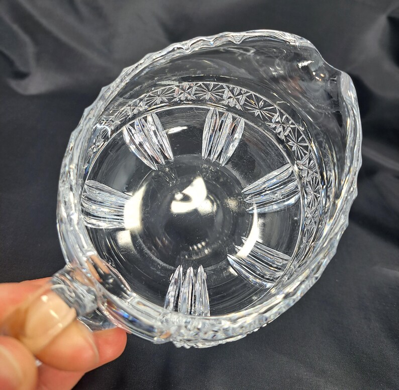 Op de afbeelding: Een creamer-schenkkan van helder glas met een decoratief geslepen patroon rond de bovenrand en verticale gegroefde details langs de binnenkant van de bodem. De kan heeft een gebogen handvat en een kleine schenktuit aan de bovenrand.