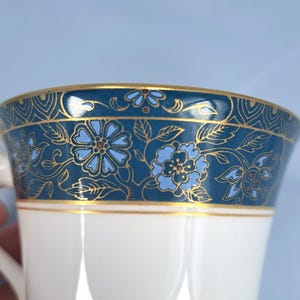 May include: A white porcelain cup featuring a wide decorative band around the top rim. The band is dark blue with a repeating pattern of light blue flowers, leaves, and intricate gold filigree lines. A thin gold line separates the band from the white base.