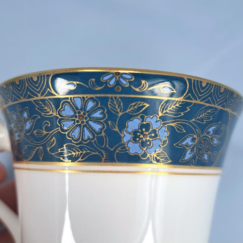 May include: A white porcelain cup featuring a wide decorative band around the top rim. The band is dark blue with a repeating pattern of light blue flowers, leaves, and intricate gold filigree lines. A thin gold line separates the band from the white base.