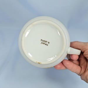 May include: White ceramic teacup with a handle, viewed from the bottom. The text "MADE IN JAPAN" is printed in black. The cup has a simple, classic design, suitable for serving tea or coffee.