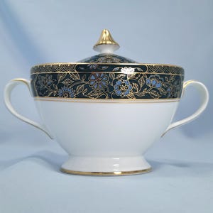 May include: A white porcelain sugar bowl with two handles and a matching lid featuring a gold finial. The upper rim is decorated with a dark teal band, intricate gold floral patterns, and light blue flower accents with gold trim edges.