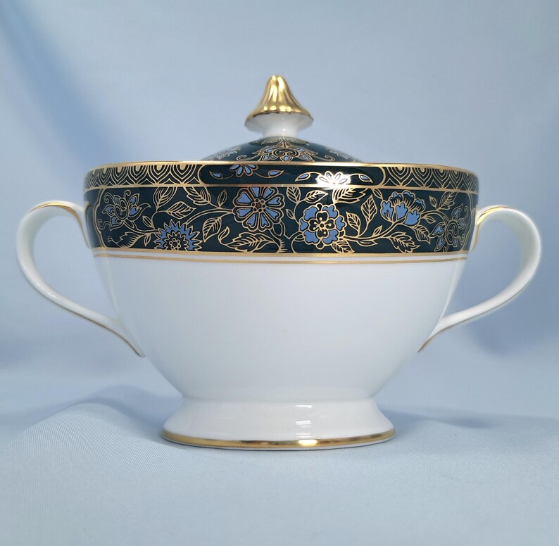 May include: A white porcelain sugar bowl with two handles and a matching lid featuring a gold finial. The upper rim is decorated with a dark teal band, intricate gold floral patterns, and light blue flower accents with gold trim edges.