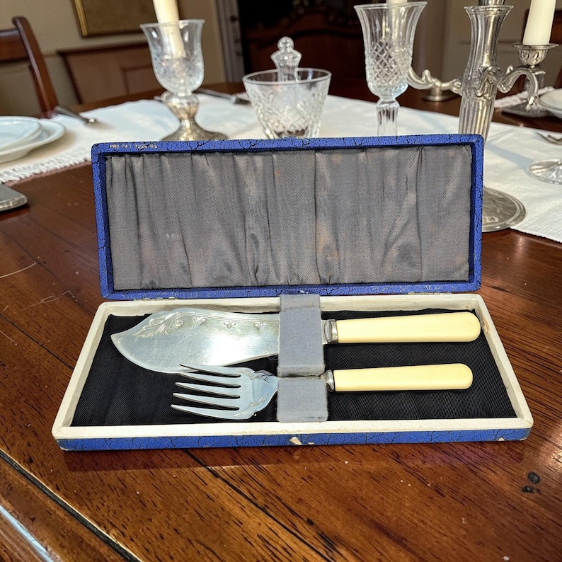 May include: A vintage carving set in a blue box. The set includes a large carving knife and a fork with cream-colored handles. The knife blade has a decorative pattern. The box is lined with black fabric.