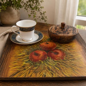 May include: A square wooden serving tray featuring a painted design of three red apples nestled in yellow straw. A white porcelain teacup with a blue and gold rim sits on the tray next to a small, round, dark brown wooden lidded container.