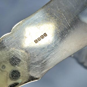 May include: Close-up of a silver-colored metal object with the letters "EPNS" stamped on it. The object has a curved, ornate design with a scalloped edge. The surface shows signs of wear.