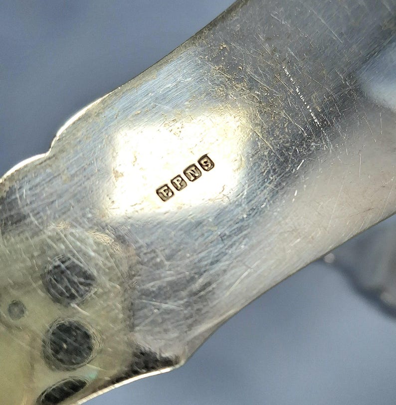 May include: Close-up of a silver-colored metal object with the letters "EPNS" stamped on it. The object has a curved, ornate design with a scalloped edge. The surface shows signs of wear.