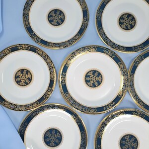 Op de afbeelding: Een set van zeven ronde porseleinen borden met een wit midden en een cirkelvormig donkerblauw en goudkleurig bloemenmedaillon. Elk bord heeft een brede donkerblauwe rand versierd met ingewikkelde gouden filigraanpatronen en een gouden rand.