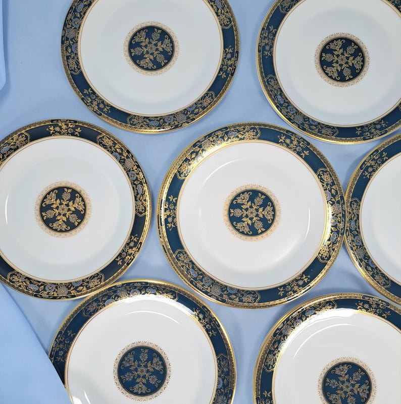 Op de afbeelding: Een set van zeven ronde porseleinen borden met een wit midden en een cirkelvormig donkerblauw en goudkleurig bloemenmedaillon. Elk bord heeft een brede donkerblauwe rand versierd met ingewikkelde gouden filigraanpatronen en een gouden rand.