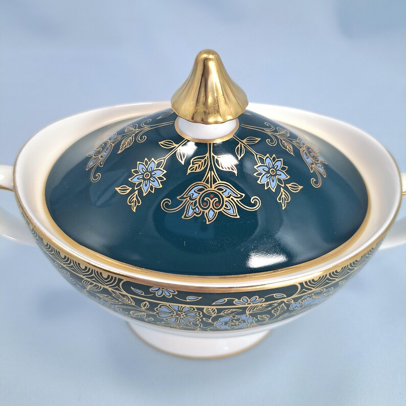 May include: A porcelain sugar bowl with a lid featuring a deep teal background, gold trim, and a repeating floral pattern in light blue and gold. The lid is topped with a gold-colored finial, and the bowl has two side handles and a pedestal base.