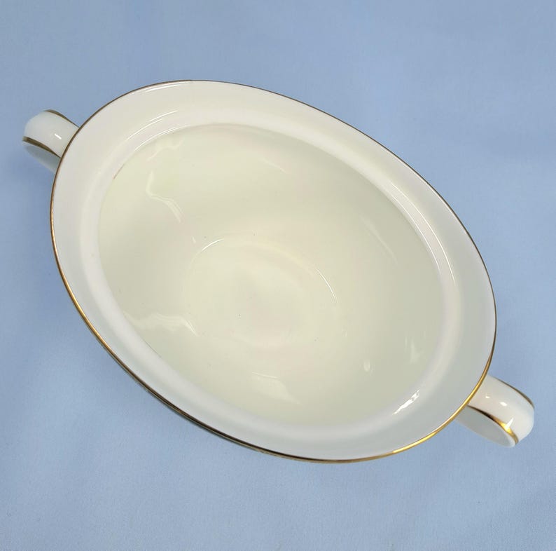 May include: An oval-shaped white porcelain serving bowl with two side handles. The rim of the bowl and the edges of the handles are accented with a thin, metallic gold trim. The interior of the bowl is smooth, glossy, and plain white.
