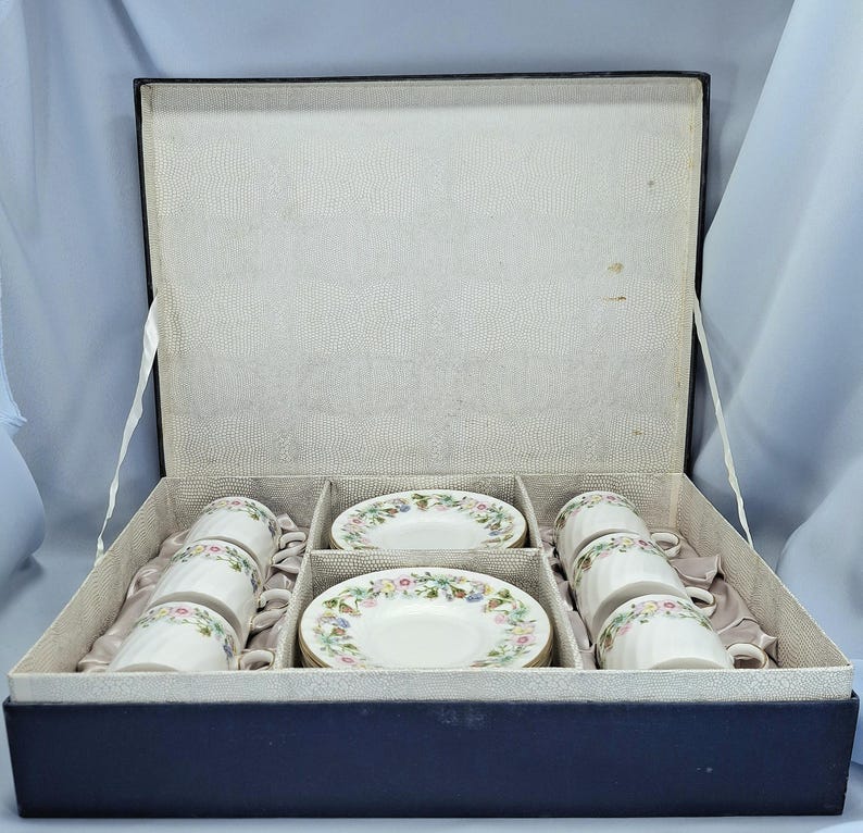 Peut inclure: Un service &agrave; th&eacute; vintage dans une bo&icirc;te bleu marine. L'ensemble comprend six tasses et soucoupes blanches, chacune d&eacute;cor&eacute;e d'un motif floral rose, jaune et bleu. L'int&eacute;rieur de la bo&icirc;te est gris clair.