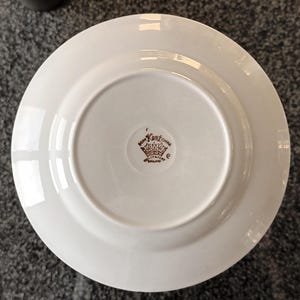 May include: White ceramic plate with a brown Kent China logo on the bottom. The logo includes a crown and the words "Kent China" and "Staffordshire England". The plate has a glossy finish.