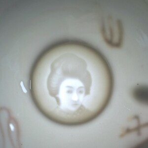 May include: A white ceramic bowl featuring a portrait of a woman with a traditional hairstyle within a circular frame. Faint red and brown decorative elements are visible around the bowl's rim.
