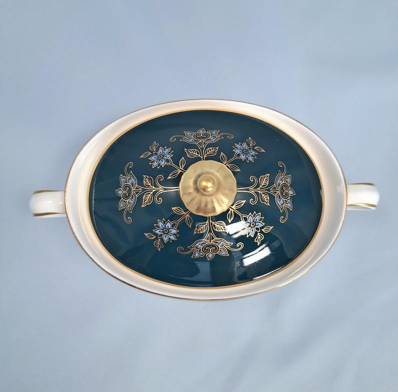 May include: An oval-shaped ceramic tureen with a dark teal lid featuring a gold and light blue floral pattern. The lid is topped with a gold-colored knob, and the tureen has white handles on both sides, set against a light blue background.