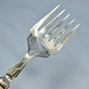 May include: Close-up of a silver fork with four tines. The fork has ornate detailing on the handle and the base of the tines. The fork is made of a shiny, reflective metal, likely silver or a silver-colored alloy. The background is a solid, light blue color.