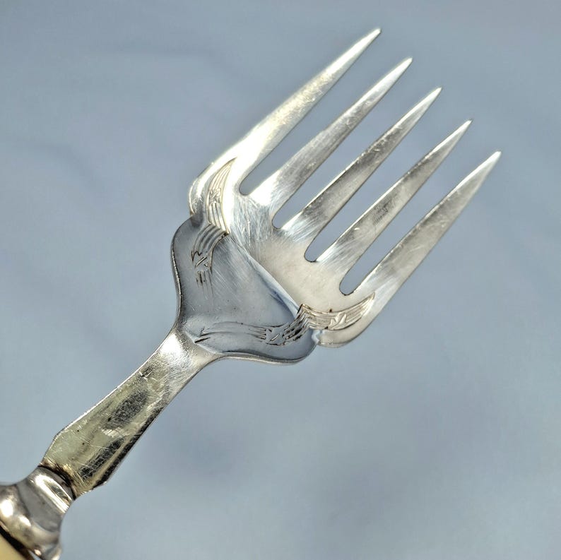 May include: Close-up of a silver fork with four tines. The fork has ornate detailing on the handle and the base of the tines. The fork is made of a shiny, reflective metal, likely silver or a silver-colored alloy. The background is a solid, light blue color.