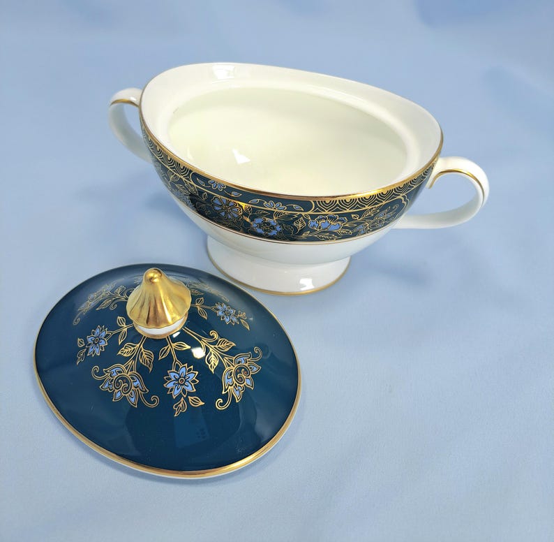 May include: A white porcelain sugar bowl with two handles and a matching lid. The bowl and lid feature a deep teal band decorated with intricate gold filigree and light blue floral patterns. The lid is topped with a pointed gold finial.