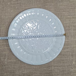 K&ouml;nnte beinhalten: Ein wei&szlig;er Keramikteller mit einem erhabenen Blumendesign in der Mitte und einem gerippten Rand. Der Teller hat einen Durchmesser von etwa 30 cm. Der Teller liegt auf einer beigen Jutefl&auml;che.