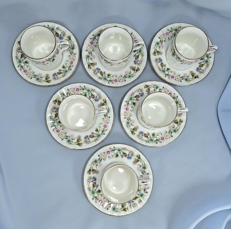 Peut inclure: Six tasses et soucoupes blanches avec un motif floral et une bordure dor&eacute;e. Les tasses et les soucoupes sont dispos&eacute;es sur une surface bleu clair. Le motif floral comprend des fleurs roses, bleues et jaunes.
