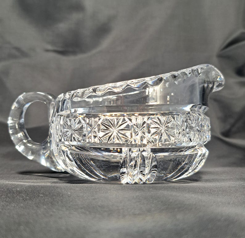 Op de afbeelding: Een creamer van helder glas met een opvallend ster- en kruispatroon rond het midden. De kan is voorzien van een gebogen handvat, een geschulpte rand en een schenktuit, met verticale gegroefde details langs de onderkant.