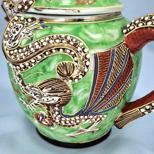 Pu&ograve; includere: Una teiera in ceramica verde con un disegno di drago. La teiera presenta un motivo di drago dettagliato in marrone, bianco e oro, con accenti rossi e blu. Il manico e il beccuccio sono rossi con finiture dorate. Il coperchio &egrave; bianco con finiture dorate.