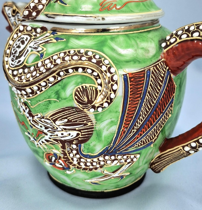 Pu&ograve; includere: Una teiera in ceramica verde con un disegno di drago. La teiera presenta un motivo di drago dettagliato in marrone, bianco e oro, con accenti rossi e blu. Il manico e il beccuccio sono rossi con finiture dorate. Il coperchio &egrave; bianco con finiture dorate.