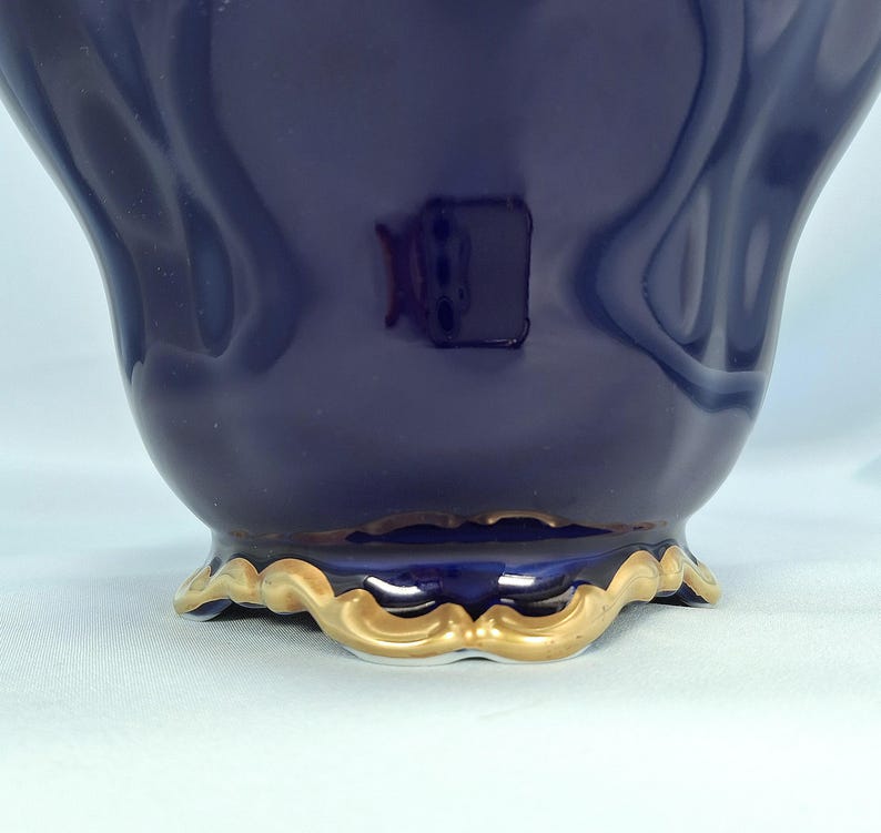 Może przedstawiać: Głęboko granatowe ceramiczne naczynie o gładkim, błyszczącym wykończeniu i zakrzywionej, falistej podstawie. Dolna krawędź naczynia jest podkreślona metalicznym złotym wykończeniem, kt&oacute;re podąża za falistym kształtem podstawy, tworząc kontrast.