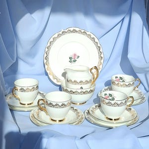May include: A vintage tea set featuring a pink rose design. The set comprises teacups, saucers, a creamer, a sugar bowl, and a serving plate. The white porcelain has gold trim and a delicate pink rose motif.