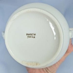 May include: White ceramic serving bowl with two handles. The base of the bowl has the text "MADE IN JAPAN" in black lettering. The bowl is round and appears to be in good condition.
