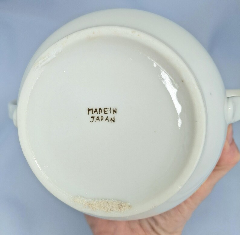 May include: White ceramic serving bowl with two handles. The base of the bowl has the text "MADE IN JAPAN" in black lettering. The bowl is round and appears to be in good condition.