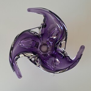 K&ouml;nnte beinhalten: Eine mundgeblasene Glasschale in tiefem Violett mit klaren Glasakzenten. Die Schale hat eine markante, dreizackige, wirbelnde sternartige Form mit geschwungenen Kanten und eine glatte, reflektierende Oberfl&auml;che, die das Licht einf&auml;ngt.