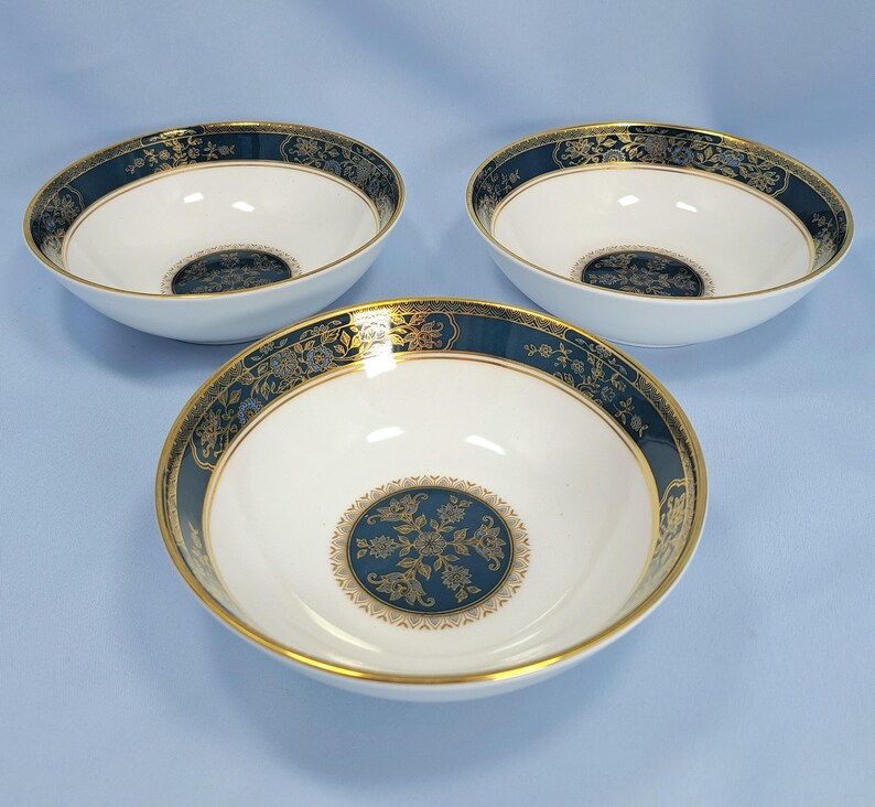 Op de afbeelding: Drie witte keramische kommen met een brede donkerblauwe rand met een goudkleurig bloemmotief en een bijpassend cirkelvormig goud en blauw bloemmotief in het midden. Elke kom heeft een dunne gouden rand aan de bovenkant en een gladde afwerking.