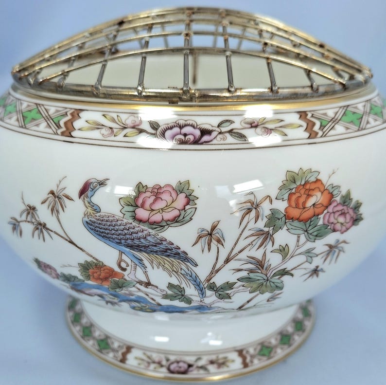 Puede incluir: Jarr&oacute;n de flores de cer&aacute;mica blanca con un dise&ntilde;o decorativo de p&aacute;jaros y flores. El jarr&oacute;n tiene una parte superior de rejilla met&aacute;lica y un borde dorado. El p&aacute;jaro es azul y las flores son rosas y naranjas.