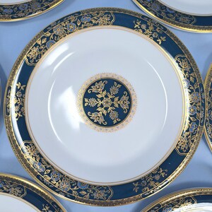 Op de afbeelding: Een wit porseleinen bord met een brede donkerblauwe rand versierd met ingewikkelde gouden bloemmotieven en een bijpassend rond goud en blauw bloemmedaillon in het midden, omlijst door dunne gouden lijnen langs de randen.