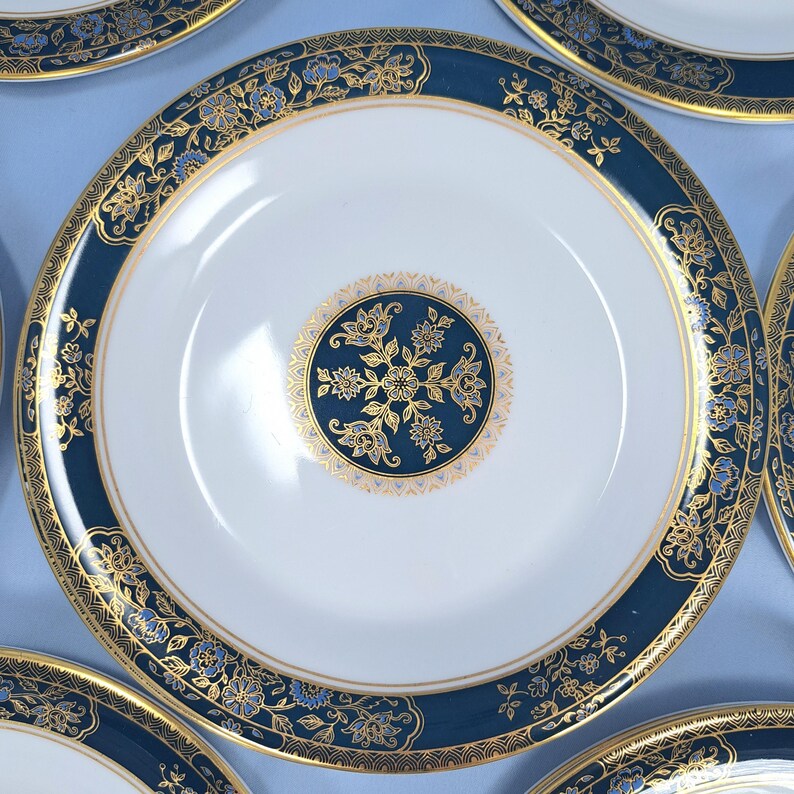 Op de afbeelding: Een wit porseleinen bord met een brede donkerblauwe rand versierd met ingewikkelde gouden bloemmotieven en een bijpassend rond goud en blauw bloemmedaillon in het midden, omlijst door dunne gouden lijnen langs de randen.