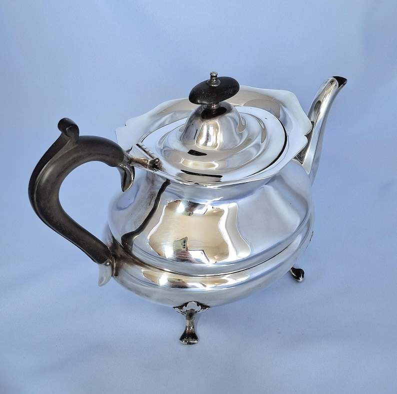K&ouml;nnte beinhalten: Eine polierte silberne Teekanne mit dunklem Griff und Deckelknopf. Die Teekanne hat einen gebogenen Ausguss und steht auf drei kleinen F&uuml;&szlig;en. Der Deckel hat ein Blumendesign um den Knopf.