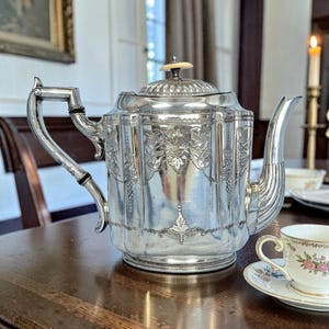 May include: A silver tea set, including a teapot with ornate floral engravings and a small teacup with a floral design. The teapot has a curved spout and a handle with black accents. The set is on a wooden table.