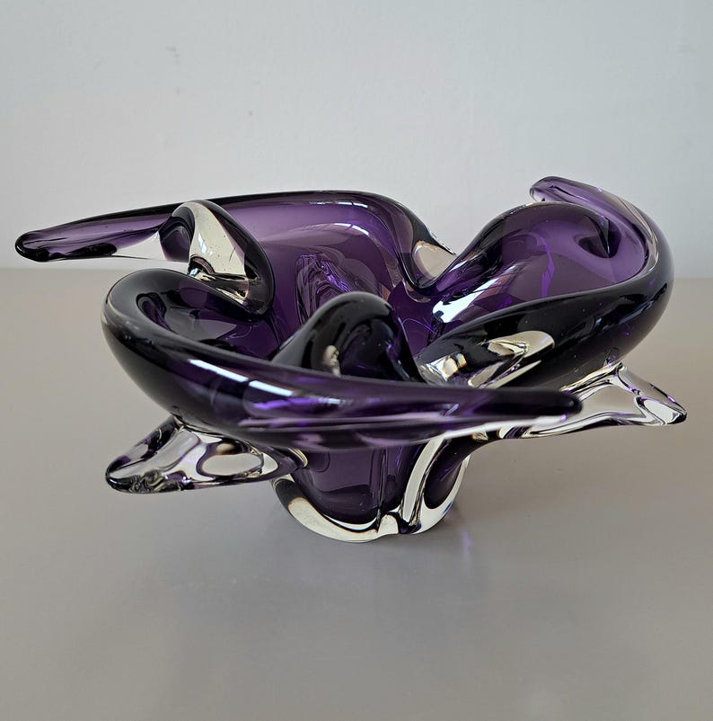 K&ouml;nnte beinhalten: Eine tiefviolette Glasschale mit organischer, freier Form, die geschwungene, gefaltete R&auml;nder sowie klare Glasakzente am Boden und am Rand aufweist. Die Schale hat eine glatte, reflektierende Oberfl&auml;che mit unterschiedlichen Transparenzgraden im Glas.
