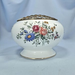 Puede incluir: Un jarr&oacute;n de flores de cer&aacute;mica blanca con borde dorado y rejilla met&aacute;lica. El jarr&oacute;n est&aacute; decorado con un estampado floral con flores rosas, azules y amarillas con hojas verdes. El jarr&oacute;n tiene unos 15 cm de di&aacute;metro.