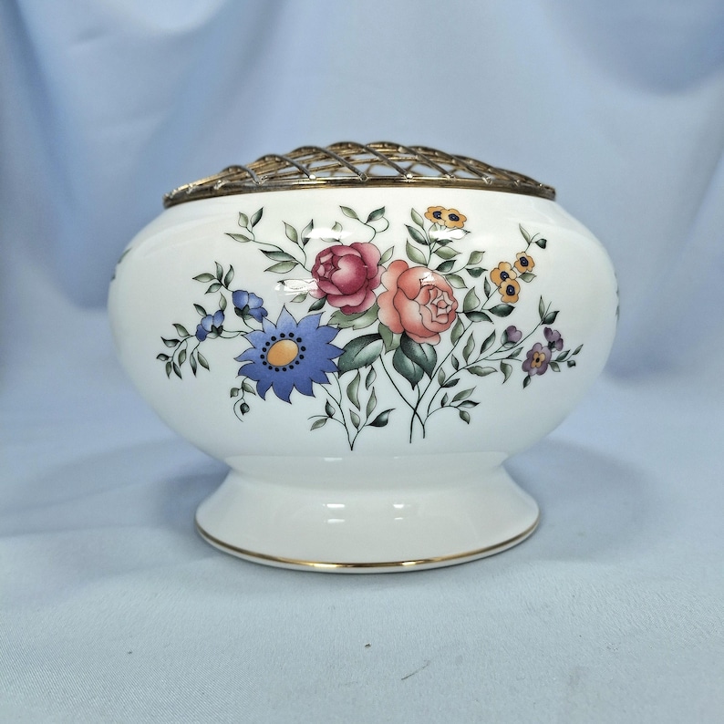 Puede incluir: Un jarr&oacute;n de flores de cer&aacute;mica blanca con borde dorado y rejilla met&aacute;lica. El jarr&oacute;n est&aacute; decorado con un estampado floral con flores rosas, azules y amarillas con hojas verdes. El jarr&oacute;n tiene unos 15 cm de di&aacute;metro.