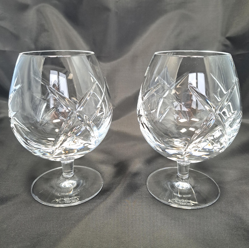 Puede incluir: Un par de copas de brandy de cristal transparente con un patr&oacute;n de corte profundo de estrella y rayado cruzado. Cada copa tiene un cuenco ancho y redondeado, un tallo corto y una base circular. El nombre Waterford est&aacute; grabado en la base.