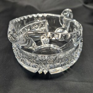 Op de afbeelding: Een asbak van helder glas met een ronde vorm, een geschulpte rand en een opvallende schenktuit. Het buitenoppervlak is versierd met een herhalend ster- en diamantpatroon, terwijl de binnenkant glad en transparant blijft.