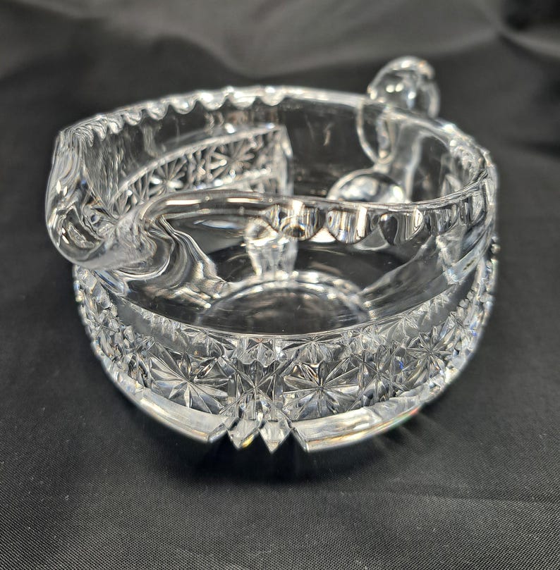 Op de afbeelding: Een asbak van helder glas met een ronde vorm, een geschulpte rand en een opvallende schenktuit. Het buitenoppervlak is versierd met een herhalend ster- en diamantpatroon, terwijl de binnenkant glad en transparant blijft.