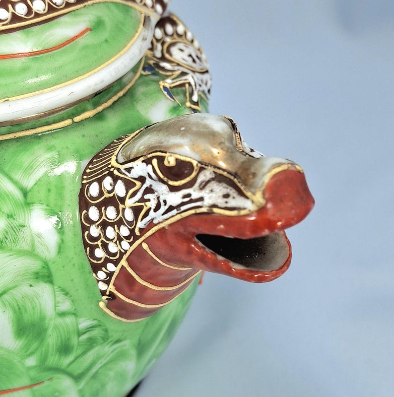 Pu&ograve; includere: Primo piano di una teiera in ceramica verde con un beccuccio a forma di testa di drago. La bocca del drago &egrave; rossa, con dettagli dorati e bianchi. La teiera ha un motivo floreale e finiture dorate.