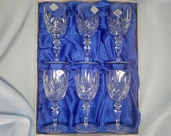 May include: A set of six clear crystal wine glasses arranged in two rows of three inside a rectangular box lined with blue satin fabric. Each glass features a faceted diamond-cut pattern on the bowl, a short stem, and a starburst base design.