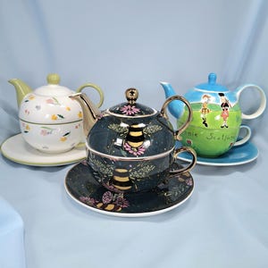 May include: Three ceramic tea sets, each consisting of a teapot stacked on a teacup with a matching saucer. One set is white with floral patterns, one is dark green with bumblebee motifs, and one is blue and green with Bonnie Scotland text.